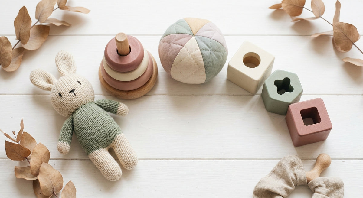 A flat lay of soft plush toys and wooden educational toys arranged together on a white wooden surface in warm, pastel tones.